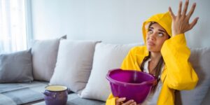 woman in raincoat holding bucket to catch ceiling leaking water